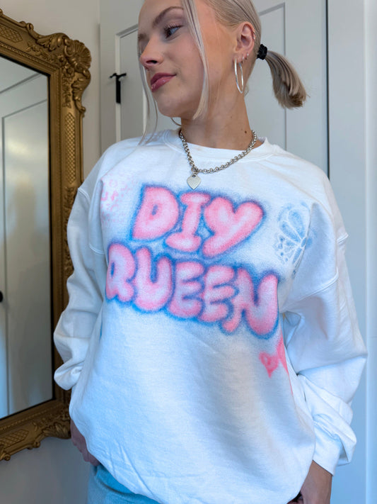 Woman standing wearing white hoodie with pink and blue airbrushed wording that reads DIY QUEEN