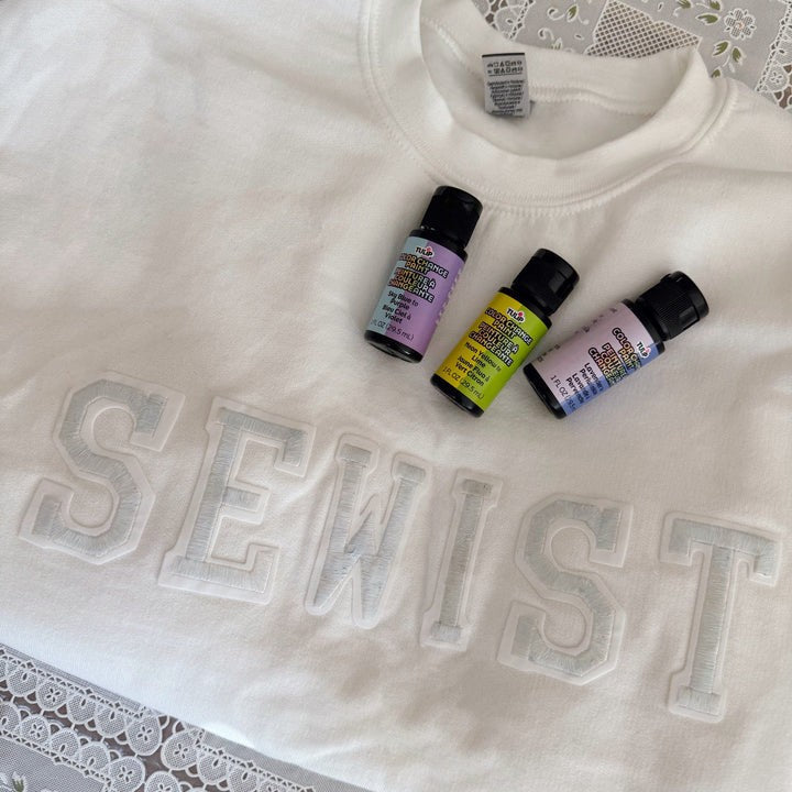 White sweatshirt with raised 'SEWIST' letters, displayed with three bottles of Tulip Soft Fabric Paint in pastel shades on top.