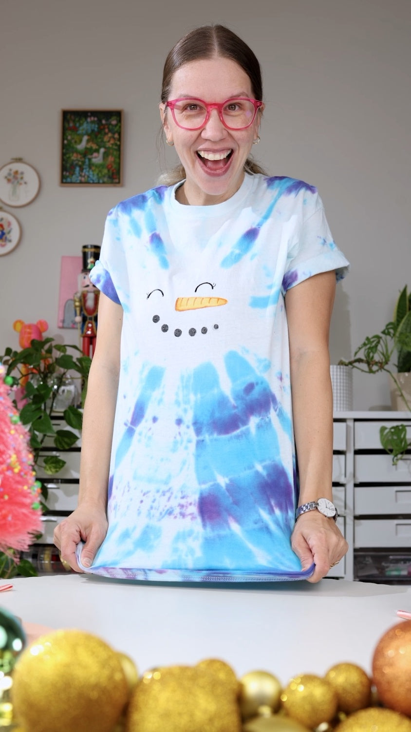 Woman standing wearing a blue bullseye tie dye shirt with painted on snowman facial features 
