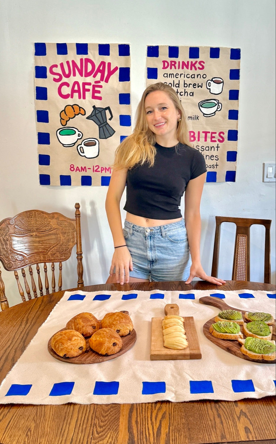 Girl standing behind a wood table with pastries on wood boards and 2 handpainted coffee shop banners on wall in background