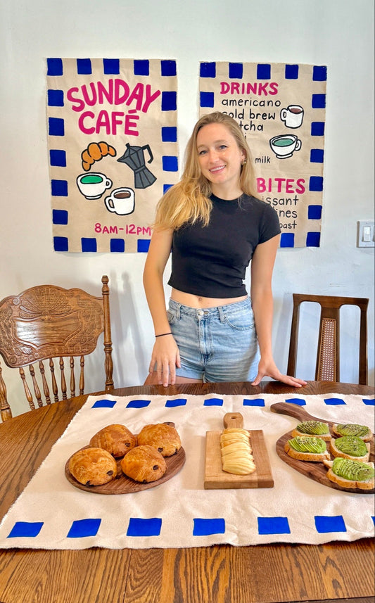 Girl standing behind a wood table with pastries on wood boards and 2 handpainted coffee shop banners on wall in background