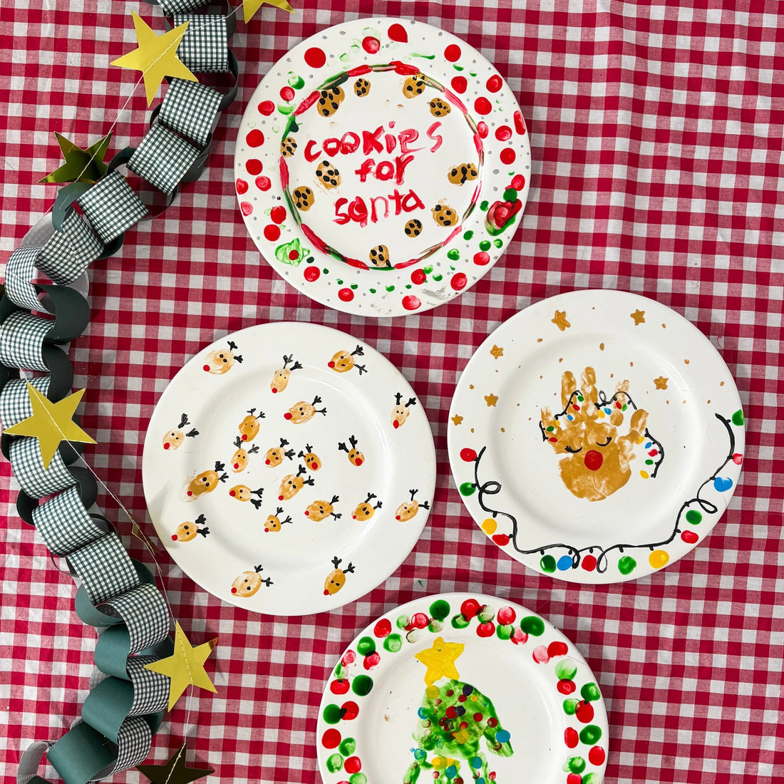 Red & white checkered tablecloth with 4 painted plates in Christmas theme and a Tulip Plate Painting Kit box on top. 