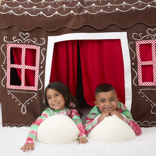 Brown fabric house decorated to look like a gingerbread house with Christmas tree on size and fake snow underneath. Two kids lie on their tummies in front of house. 