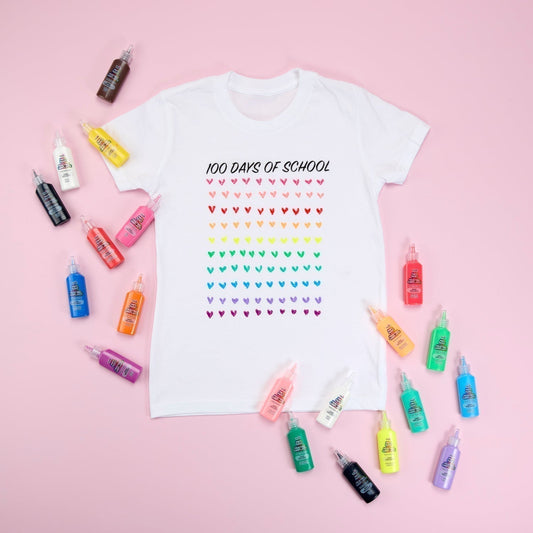White tee with "100 Days of School" printed in black at top and 100 colorful hears painted in rows. Bottles of Puff Paint surround shirt and all sits on pink background.