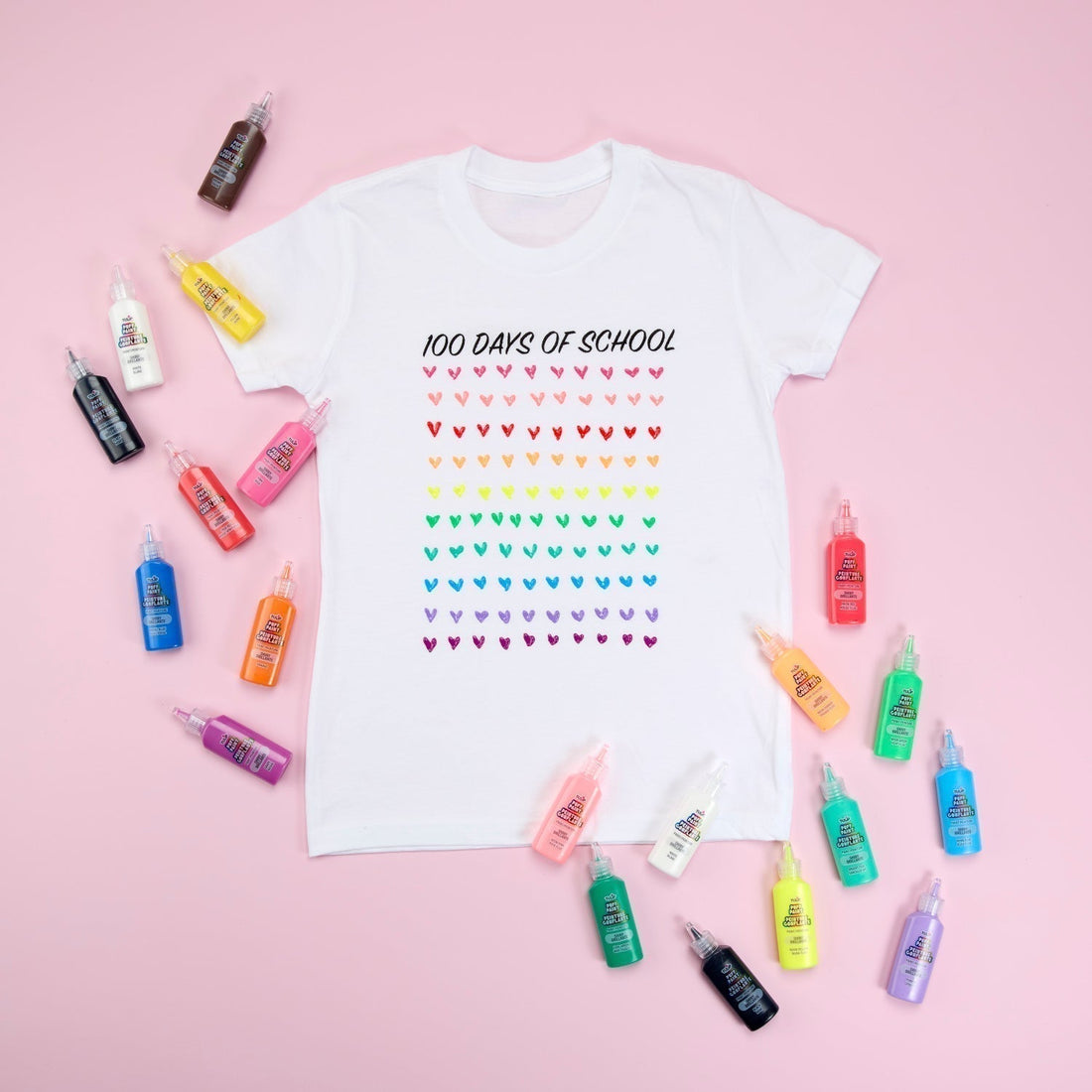 White tee with "100 Days of School" printed in black at top and 100 colorful hears painted in rows. Bottles of Puff Paint surround shirt and all sits on pink background.