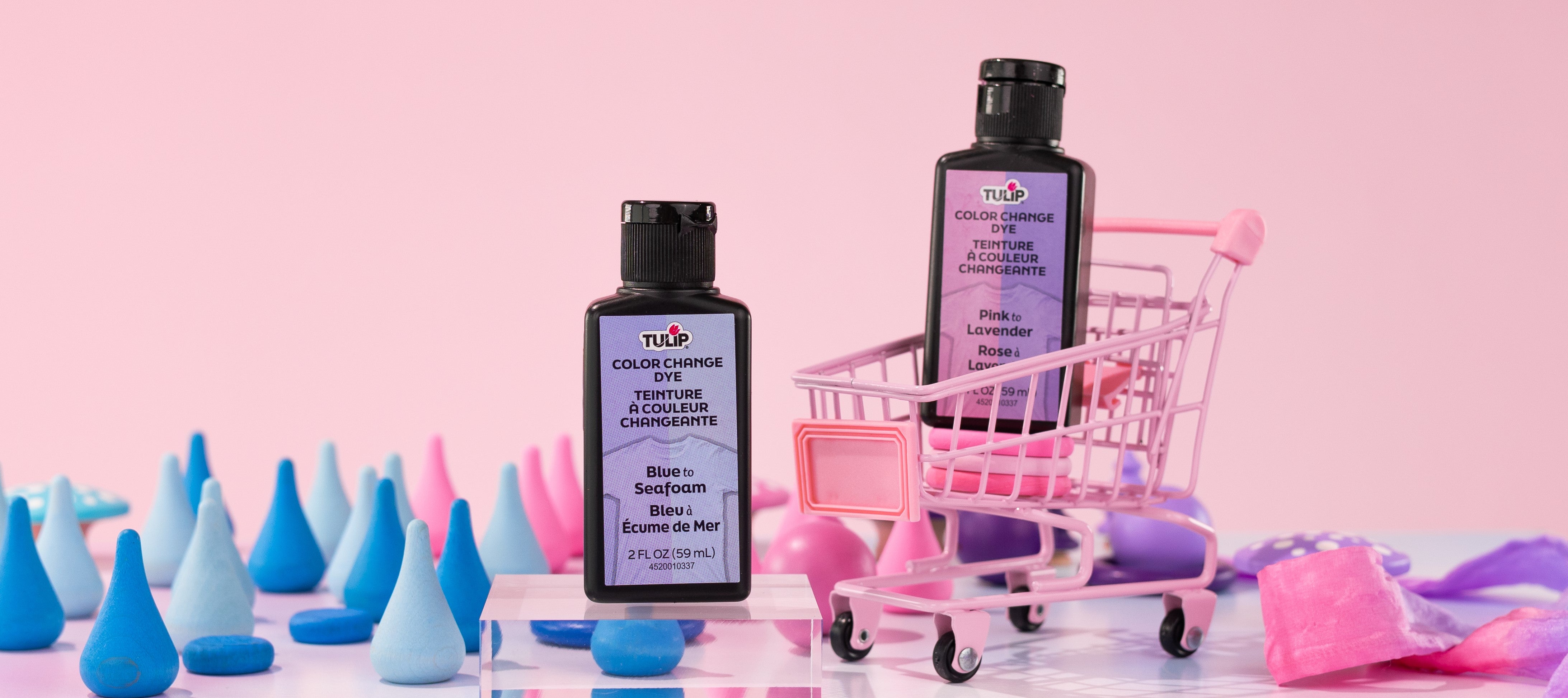 Two bottles of Tulip Color Change Dye in shades 'Blue to Seafoam' and 'Pink to Lavender' are displayed in a playful, pastel setup with colorful cone shapes and a mini pink shopping cart against a pink background.