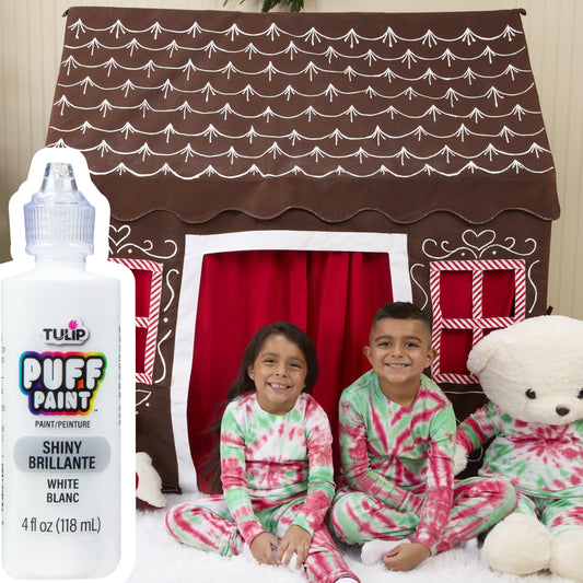 Children in pajamas with a teddy bear in front of a brown playhouse with white roof, next to a bottle of Tulip Puff Paint.